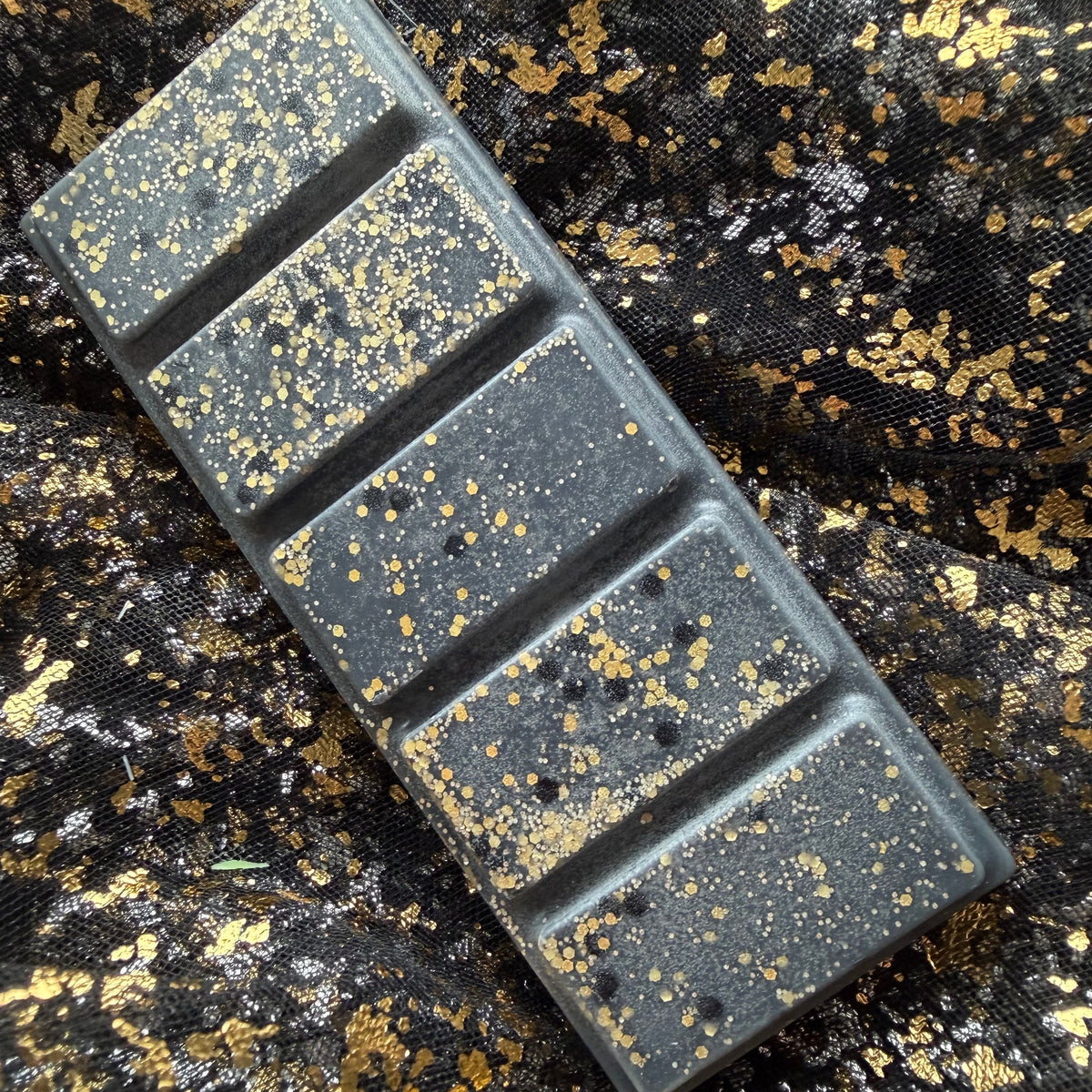 Blonde Bombshell bookish wax melt, shown sideways as a charcoal-black rectangular five-segment soy wax bar scattered with gold glitter and black circular decoration on a black-and-gold fabric background.
