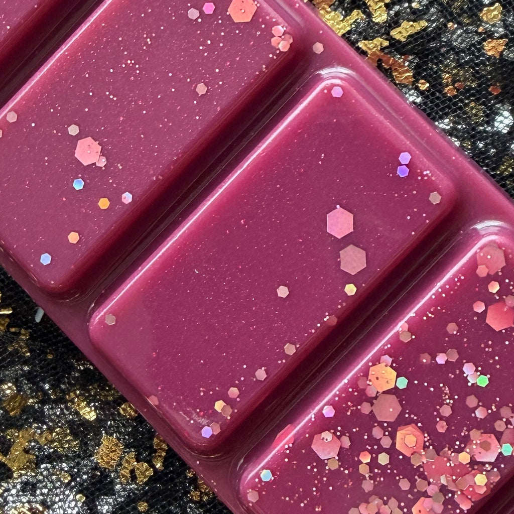 Close-up of three deep burgundy Cherry Bomb wax melt segments decorated with fine pink shimmer and pink iridescent hexagonal glitter against black-and-gold fabric.
