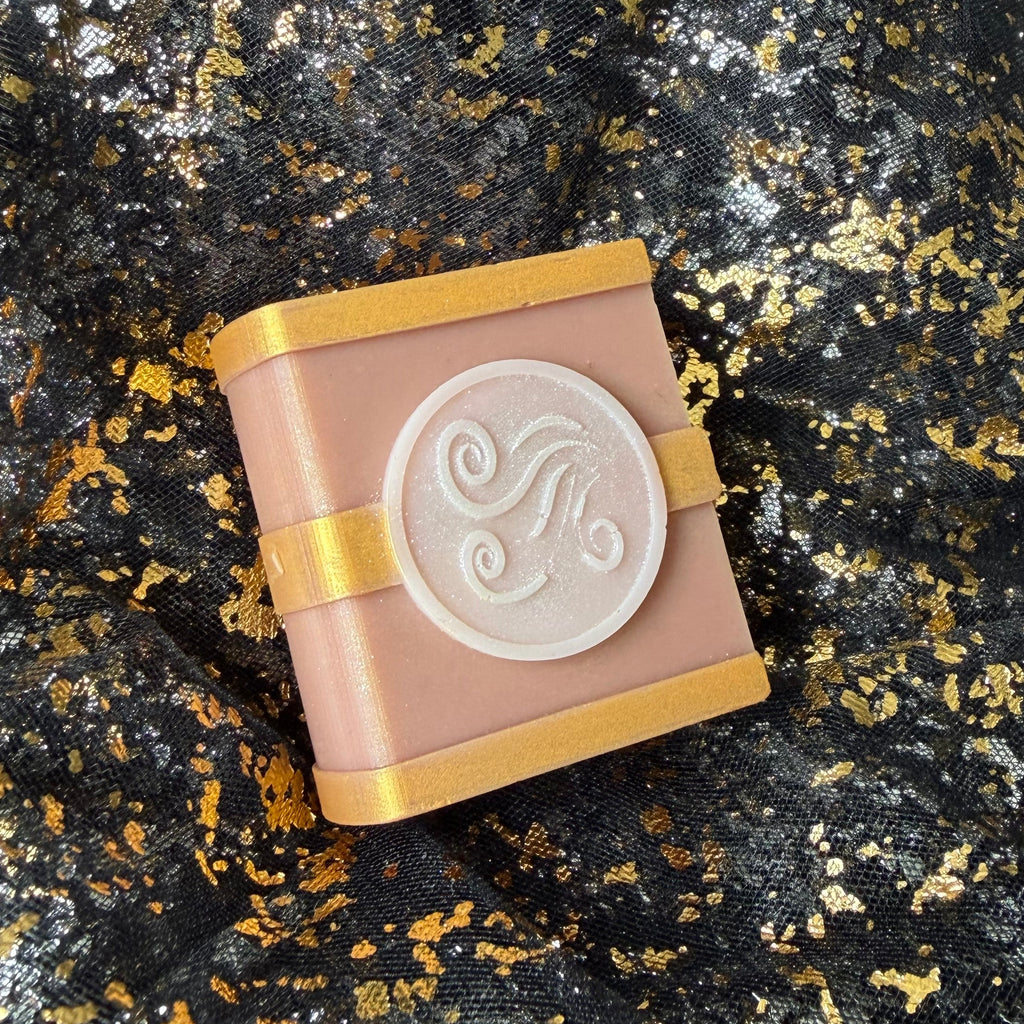 Air soy wax spellbook tilted on black and gold fabric, with its rounded spine beside a pearly wind swirl emblem and gold cover detailing.