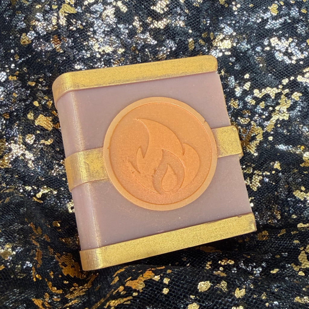 Side angled view of the Fire spellbook wax melt, with its rounded spine, gold bands and orange flame emblem visible on black and gold fabric.
