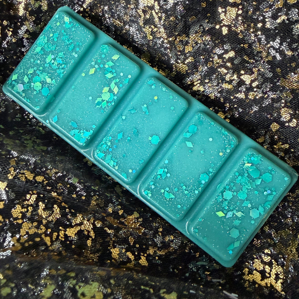 Full-length turquoise Malibu vegan soy wax melt positioned diagonally on black-and-gold fabric, showing all five segments and scattered blue-green iridescent glitter.