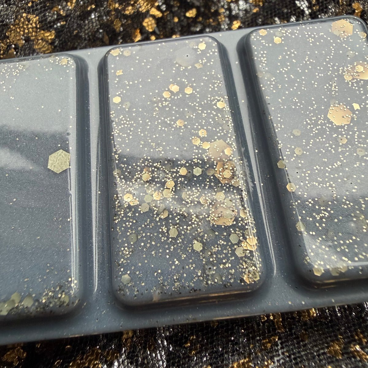 Close-up of three I Prefer the Villain wax melt segments, showing fine metallic gold glitter and larger gold hexagonal pieces against the dark charcoal wax.