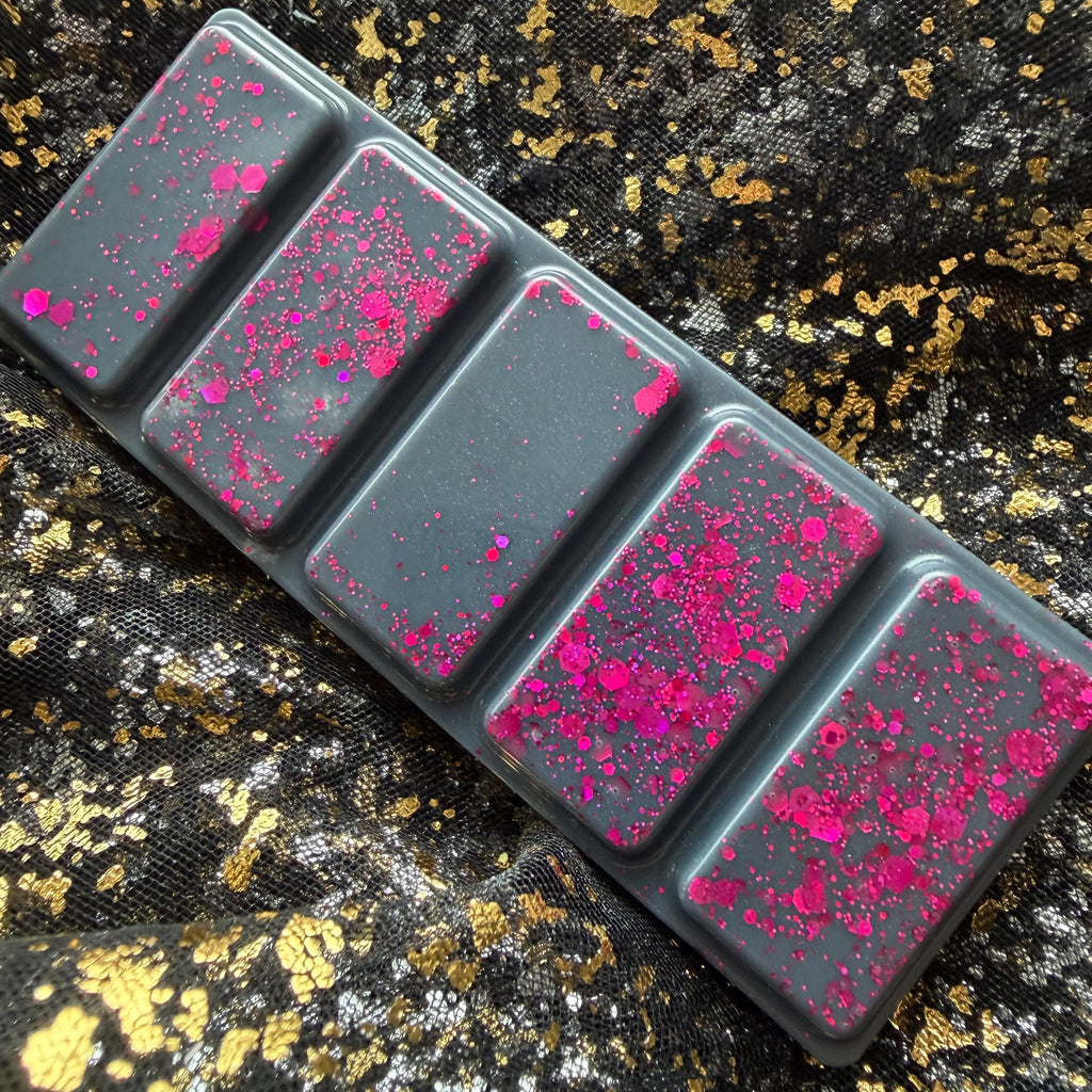 Charcoal-grey Black Beak Witch wax melt displayed diagonally on black-and-gold fabric, showing five rectangular segments decorated with vivid pink glitter.