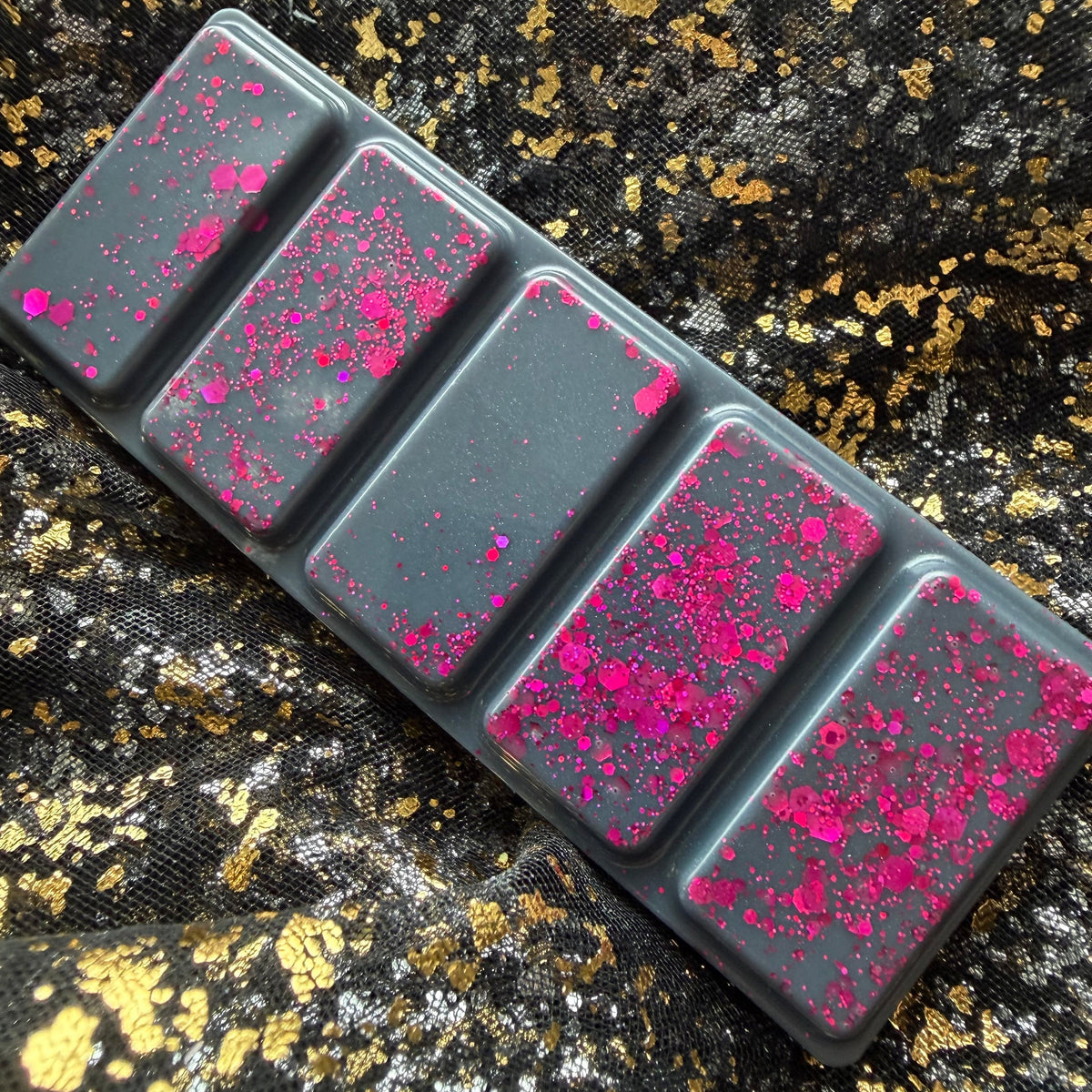 Charcoal-grey Black Beak Witch wax melt displayed diagonally on black-and-gold fabric, showing five rectangular segments decorated with vivid pink glitter.