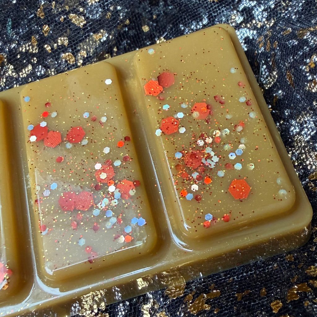 Zoomed-in view of one end segment of the A Winter in Stars Hollow Christmas-spice wax melt, showing red, gold and silver glitter scattered across the caramel-brown wax on black-and-gold fabric.