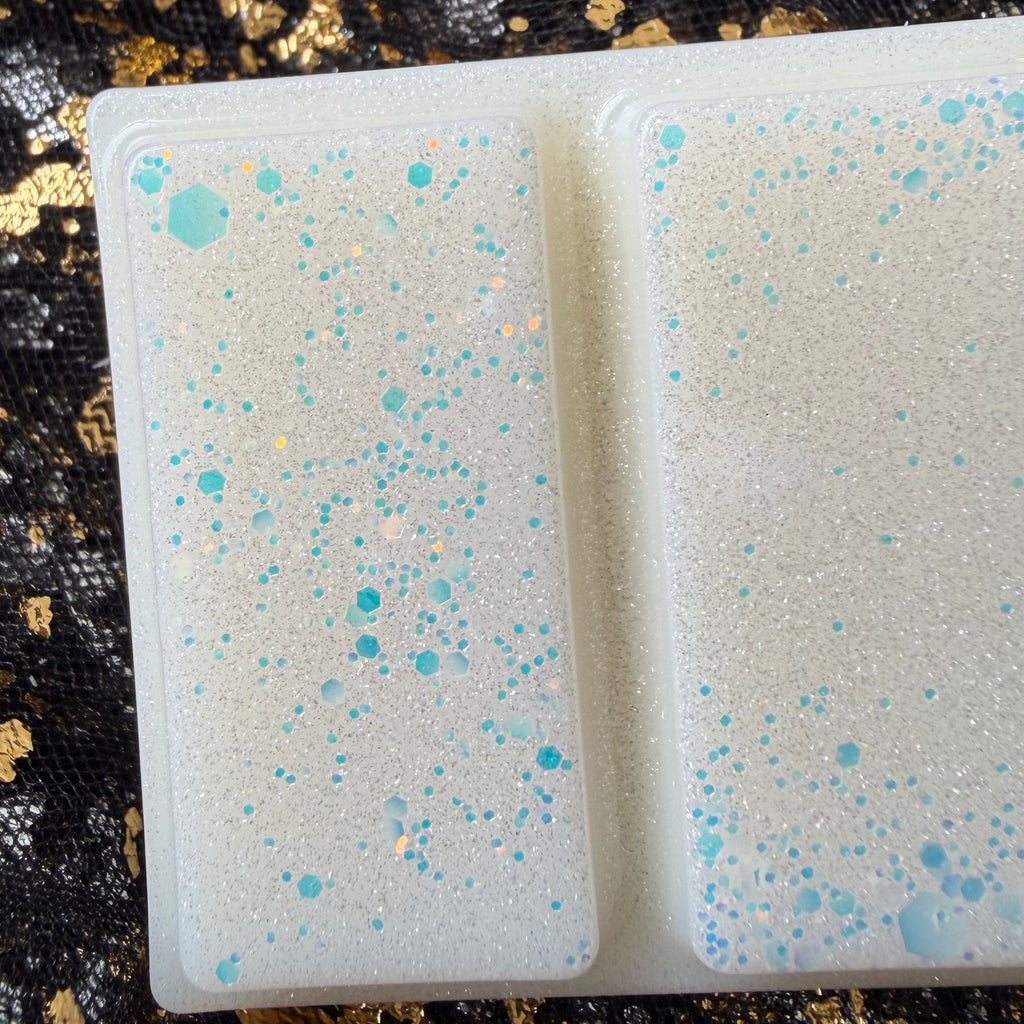 Close view of two Xavier Acrux wax melt segments, with pale blue hexagonal glitter in different sizes and fine silver shimmer across the white wax.