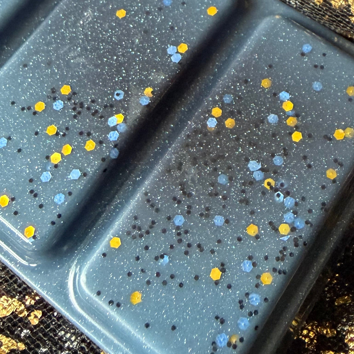 A close-up of a rectangular gray mold or tray with four raised sections, filled with glittery gray material speckled with small blue and yellow confetti, resting on a black mesh fabric with gold flecks.