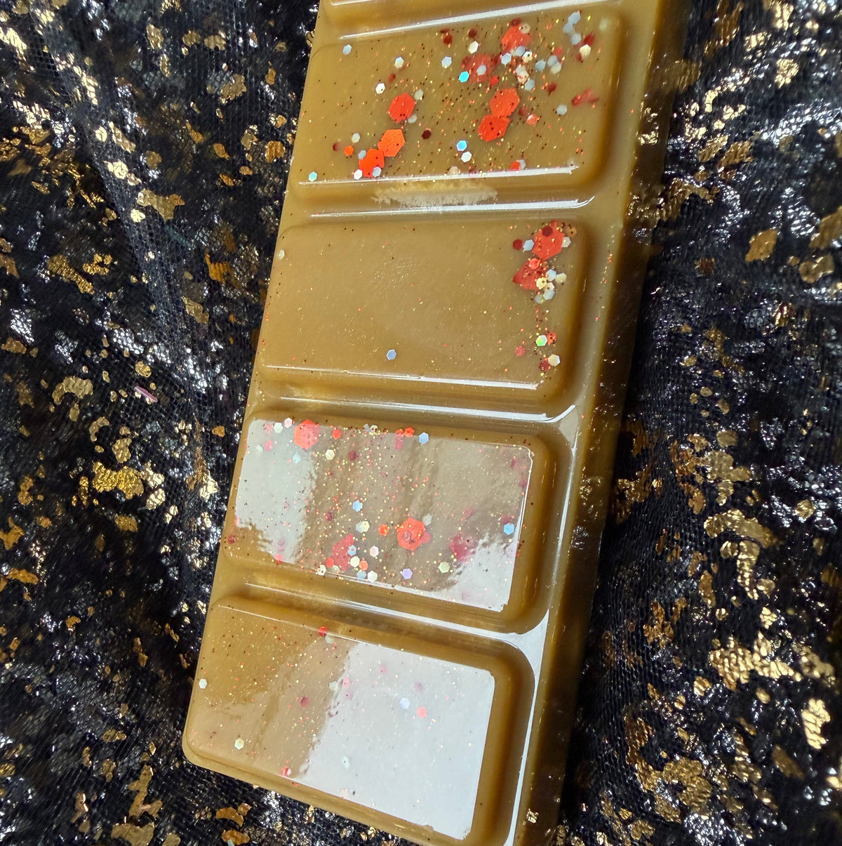 Angled close-up of the A Winter in Stars Hollow wax melt, showing caramel-brown rectangular segments decorated with red, gold and silver hexagonal glitter on a black-and-gold fabric background.