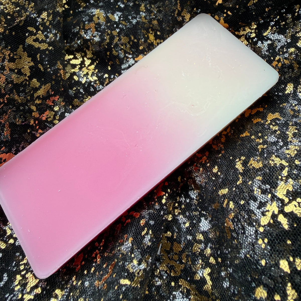 Underside of the Rosy Jam wax melt displayed diagonally on black-and-gold fabric, showing a smooth colour gradient from vivid pink into creamy white.