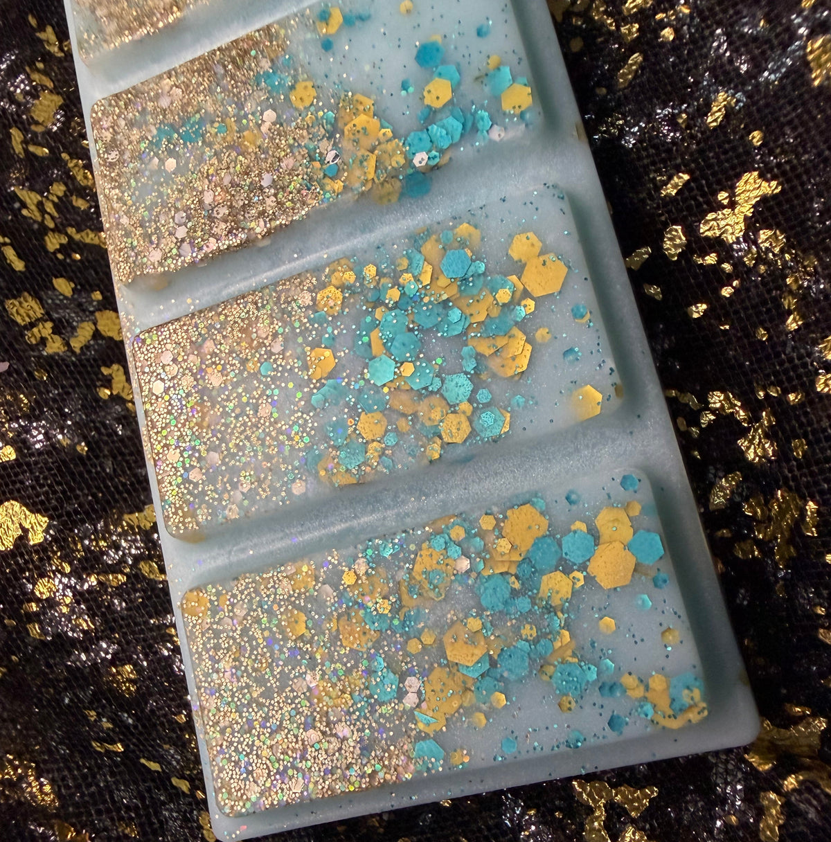 A close-up of an ocean scented soy wax melt filled with glitter and scattered gold and turquoise blue flakes, placed on a black fabric background with gold flecks.
