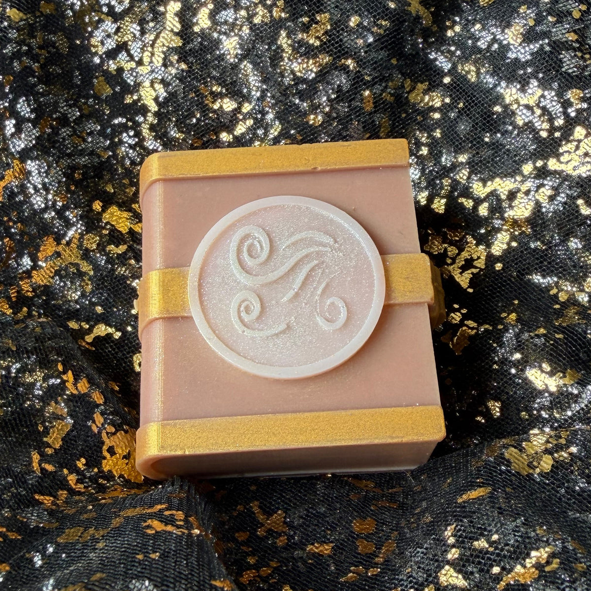 Close angled view of the Air spellbook wax melt, with a shimmering wind emblem, gold bands and the lower edge of the miniature book visible.