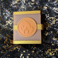 Close view of the Fire mango and vanilla scented spellbook wax melt, with an orange flame emblem between gold bands on its dusty rose cover.