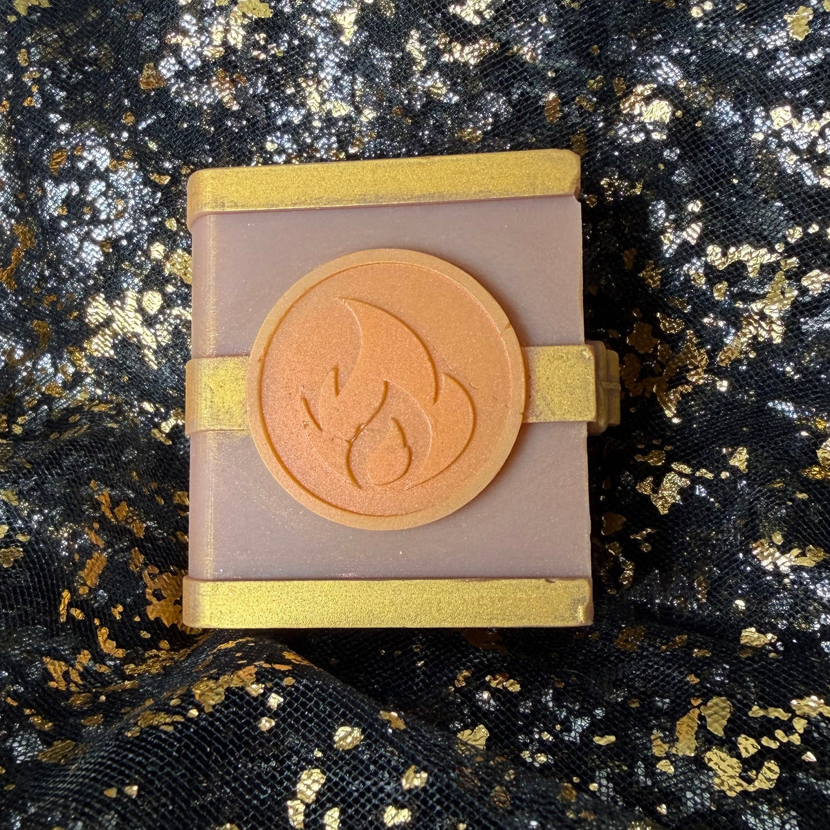 Close view of the Fire mango and vanilla scented spellbook wax melt, with an orange flame emblem between gold bands on its dusty rose cover.