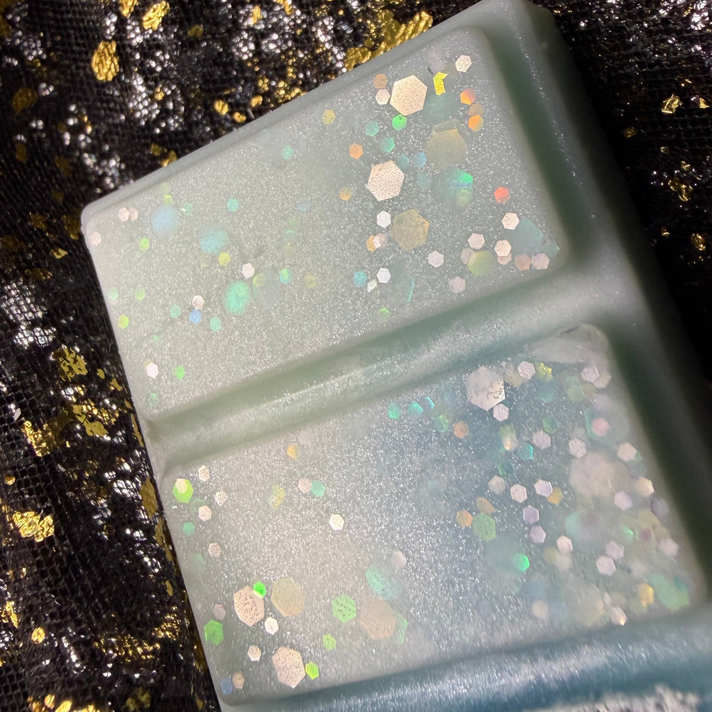 Close-up of two pale aqua Lime Basil & Mandarin wax melt segments, showing scattered silver, blue and iridescent hexagonal glitter.