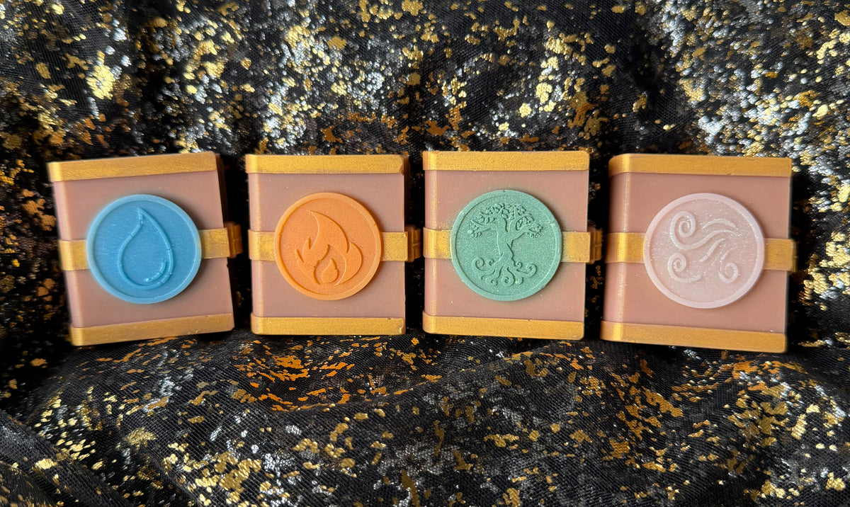 Four unwrapped Zodiac Academy soy wax spellbooks standing on black and gold fabric, with blue Water, orange Fire, mint Earth and pearly Air emblems from left to right.