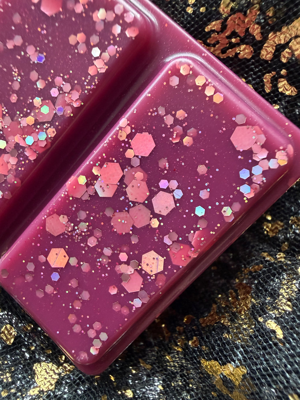 Zoomed-in view of a Cherry Bomb wax melt segment showing deep burgundy wax decorated with large pink hexagons, smaller iridescent glitter and fine shimmer.
