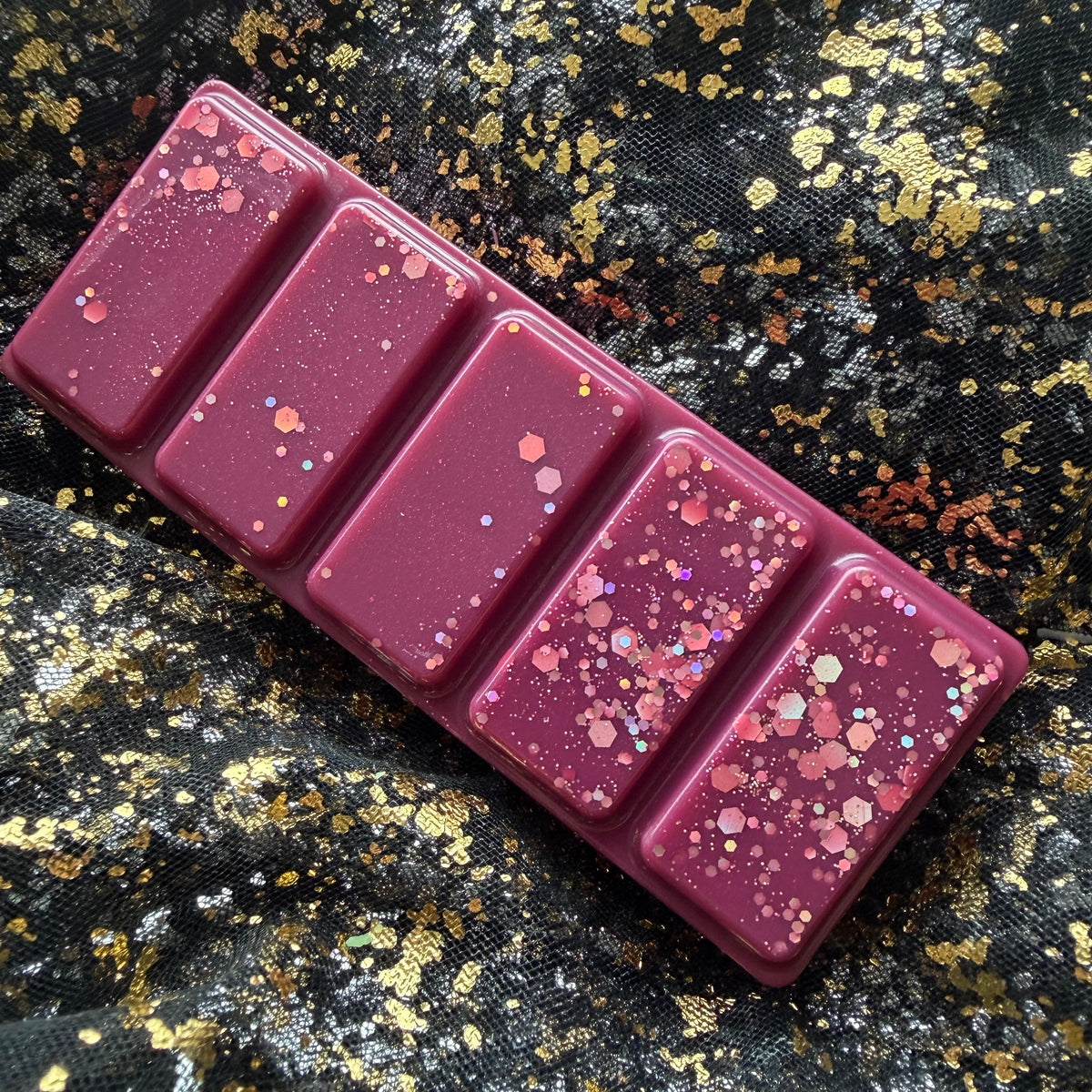 Cherry Bomb black cherry wax melt, shown sideways as a deep burgundy rectangular five-segment soy wax bar scattered with pink and iridescent hexagonal glitter on a black-and-gold fabric background.
