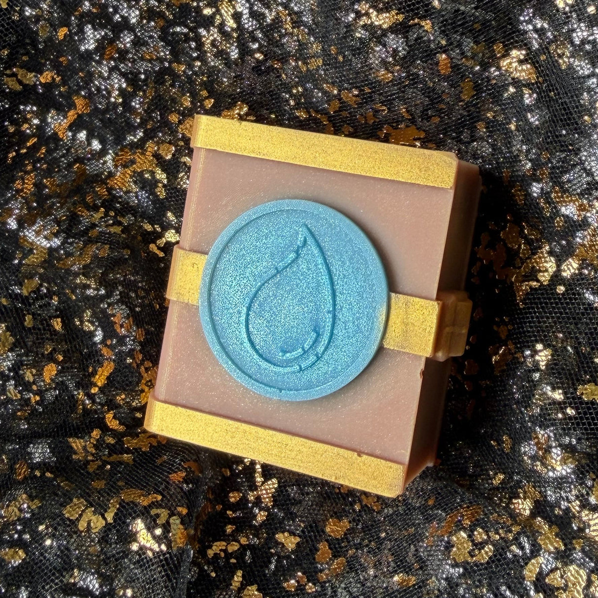 Side view of the Water spellbook wax melt, with a silvery blue droplet emblem, gold clasp and the right edge of the book visible.