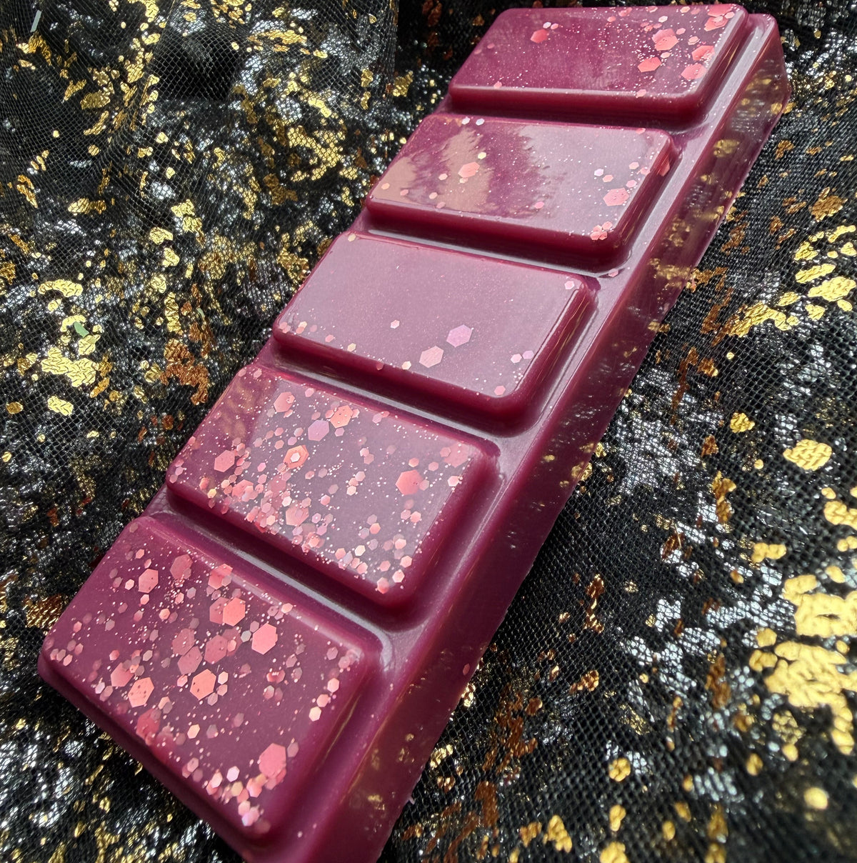 Low angled view along the raised rectangular segments of the Cherry Bomb wax melt, showing deep burgundy wax with clusters of pink iridescent glitter on black-and-gold fabric.
