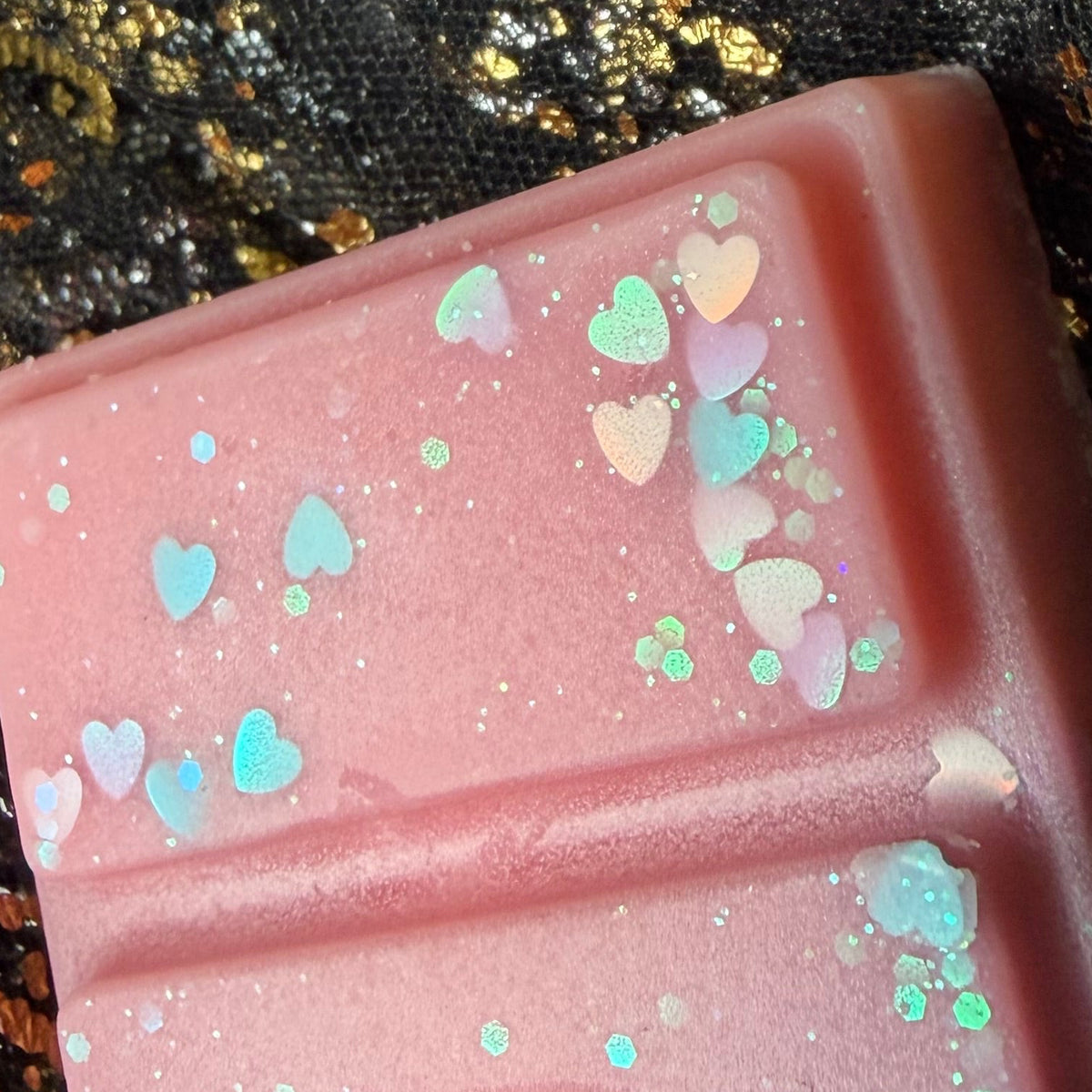 A close up of  the pink side of the black raspberry and vanilla soy wax melt decorated with pastel heart-shaped glitter and iridescent flecks, placed on a black fabric background with gold foil scattered around.