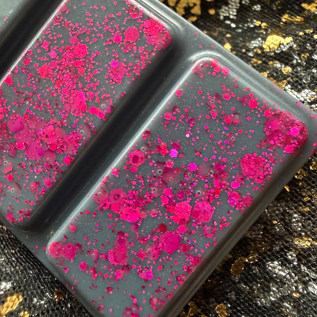 Close-up of two Black Beak Witch wax melt segments showing dense vivid pink glitter and larger pink pieces against charcoal-grey wax.