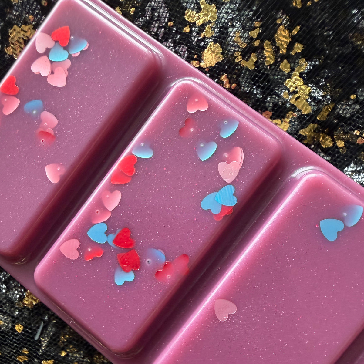 Angled close-up of three Bryce After Dark wax melt segments showing deep berry-purple wax, fine shimmer and scattered red, pink and blue heart-shaped glitter.