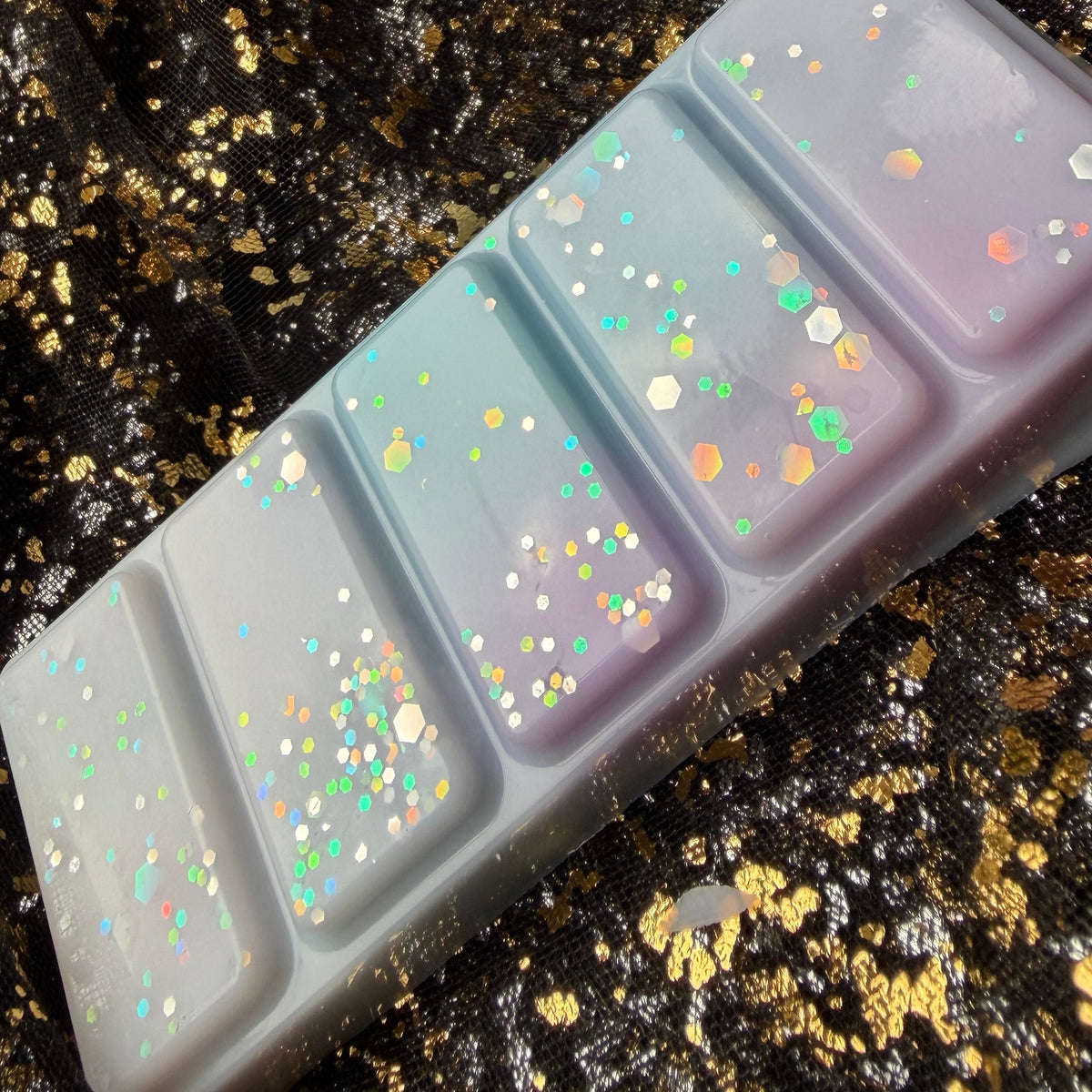 shiny spring awakening scented Spring Court soy wax melt displayed diagonally on black-and-gold fabric, showing blue, white and lilac segments decorated with iridescent glitter.