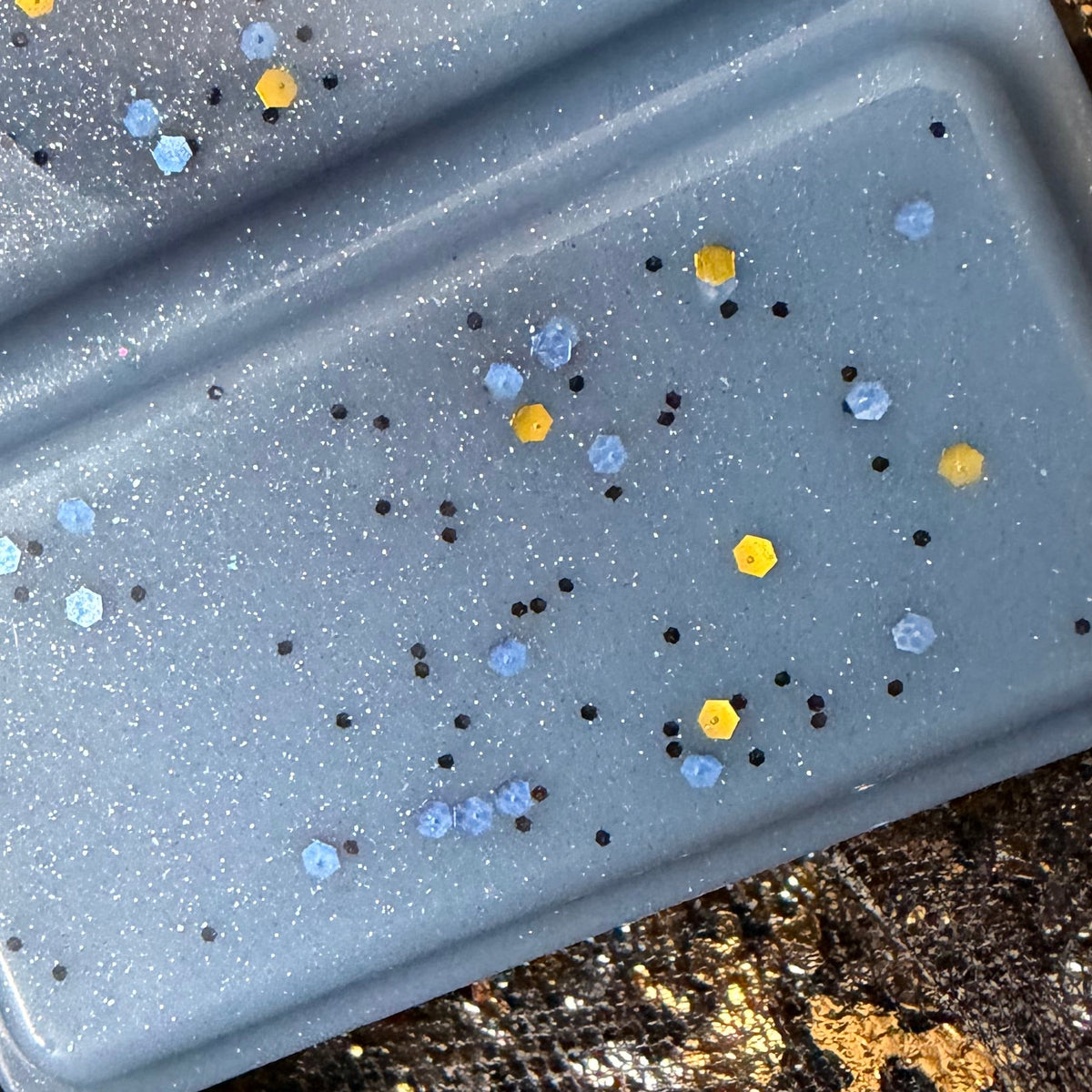 Close-up of the upper Morally Grey soy aftershave scented wax melt segments showing yellow, silver, blue and black glitter against slate-grey wax.