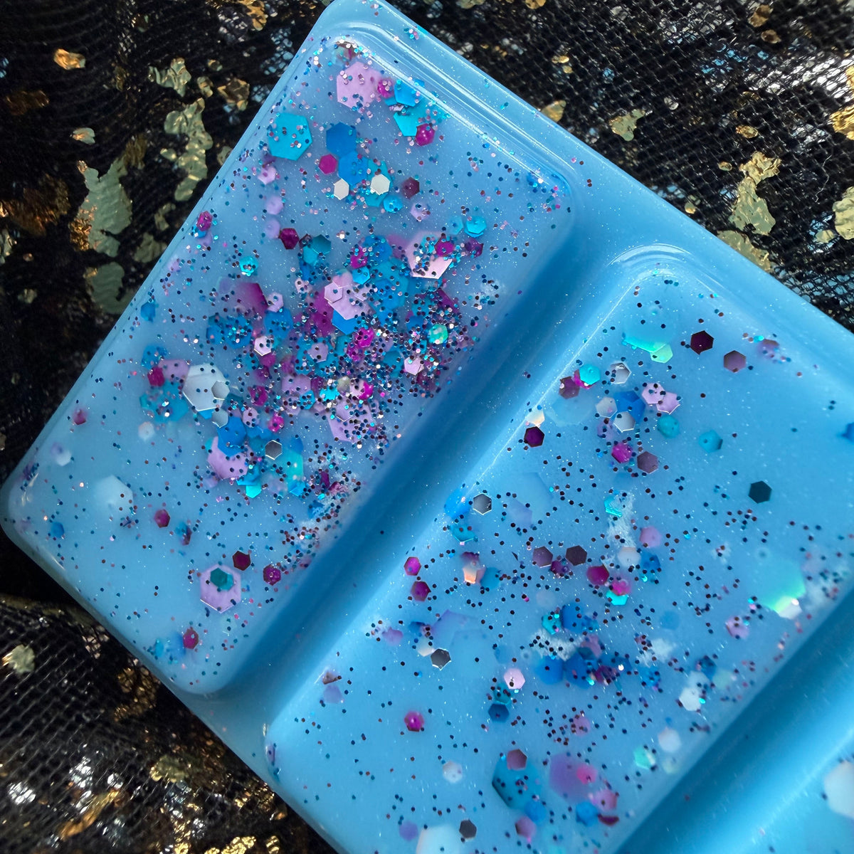 Close-up of two Starfall wax melt segments showing pink, purple, blue and silver glitter scattered across bright turquoise wax.