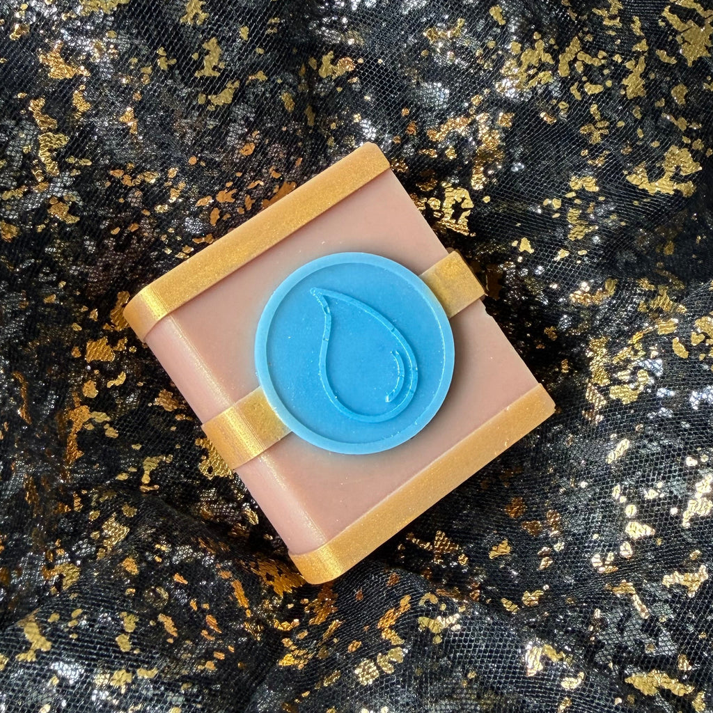 Overhead photograph of The Awakening Water wax melt, showing the full miniature spellbook with its blue emblem and gold detailing on black and gold fabric.