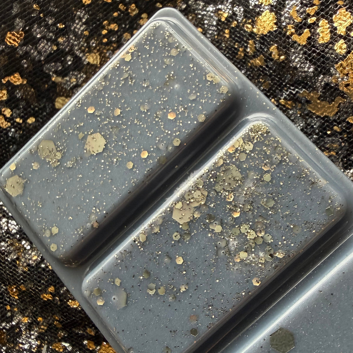 Zoomed-in photograph of the end sections of the I Prefer the Villain wax melt, showing clusters of metallic gold flecks and hexagonal glitter pieces on dark charcoal wax.