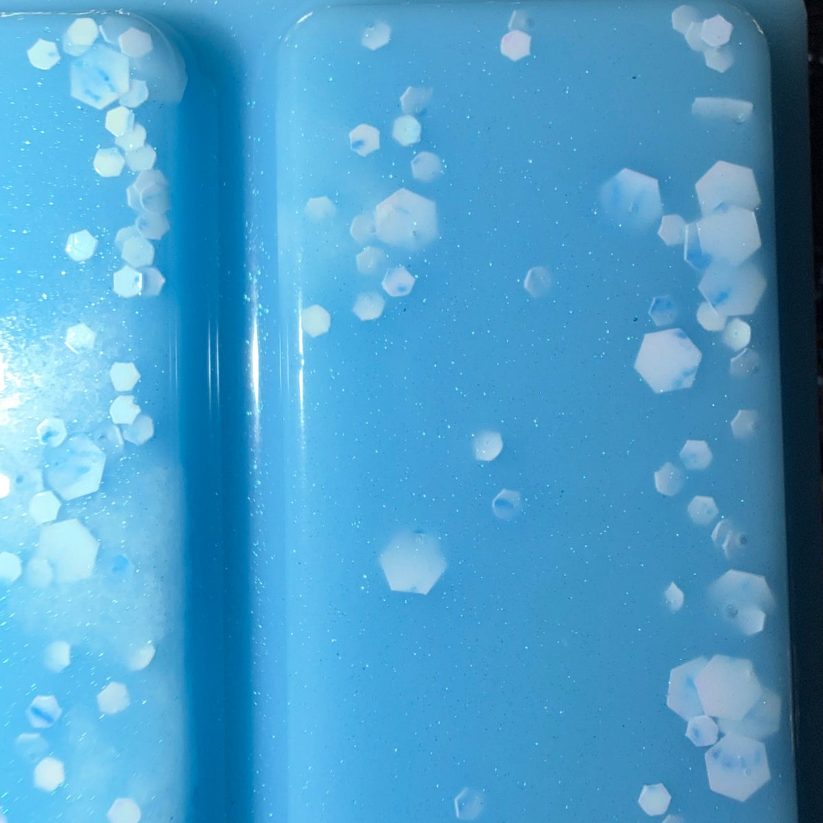 Zoomed-in photograph of The Cleaning Fairy wax melt showing white hexagonal glitter, fine silver shimmer and tiny iridescent pieces against blue wax.