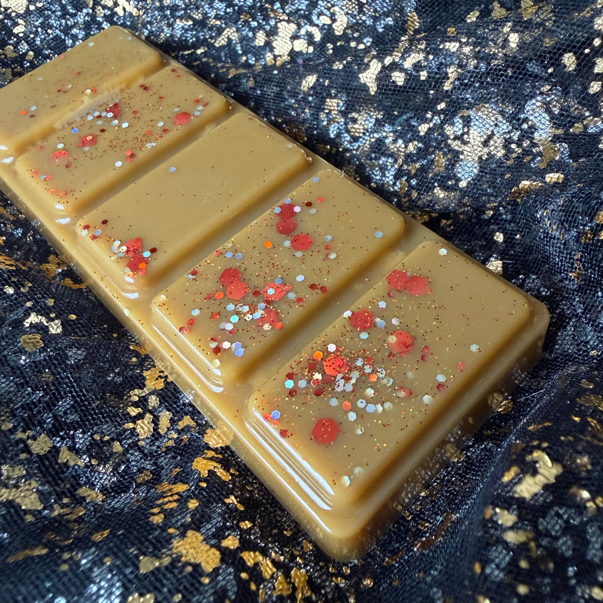 A Winter in Stars Hollow is a christmas spiced scented wax melt, shown sideways as a caramel-brown rectangular five-segment soy wax bar with red, gold and silver glitter, positioned diagonally on black-and-gold fabric.