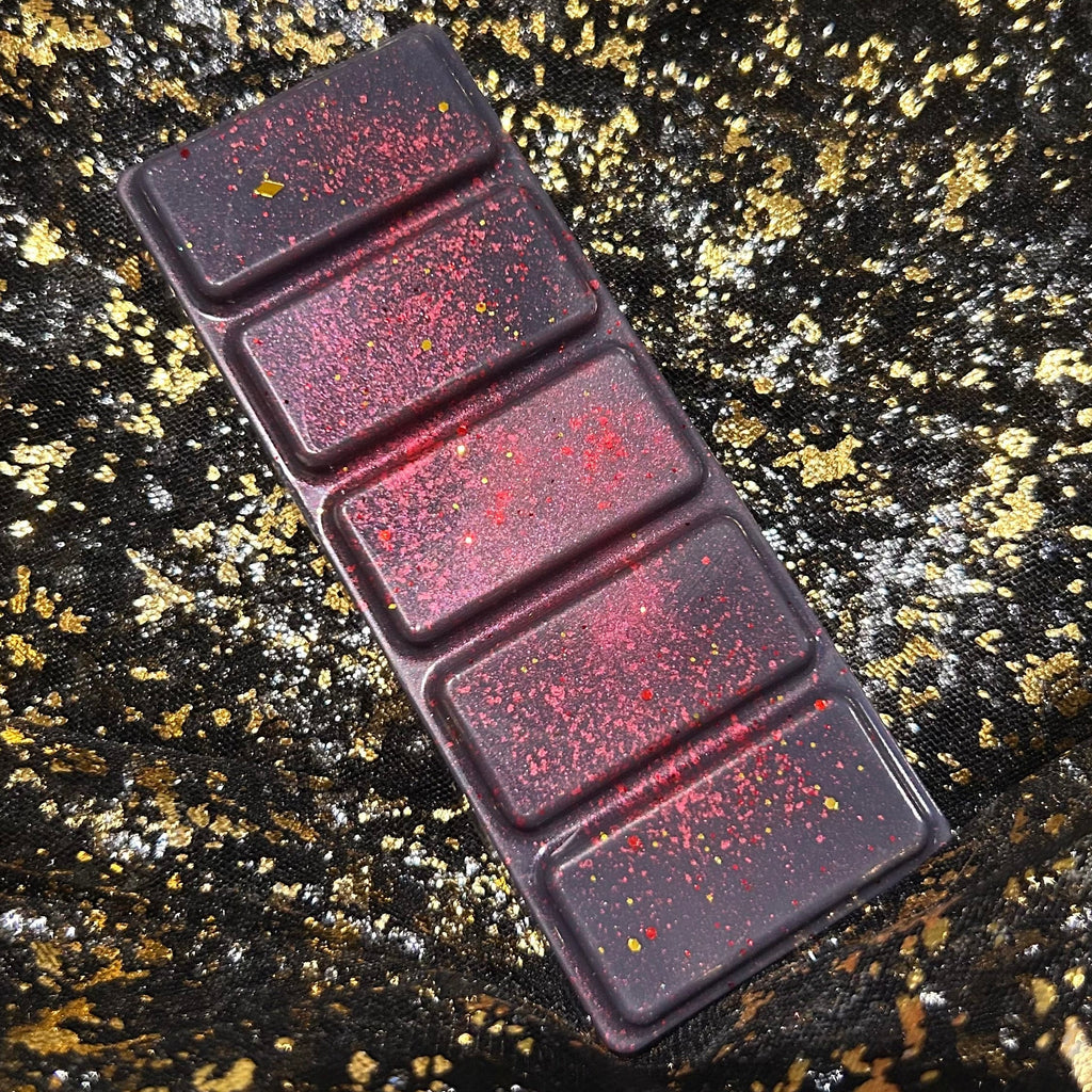 Full Tory Vega burgundy wax melt bar on folded black and gold fabric, with five rectangular segments, fine red shimmer and small gold glitter pieces.
