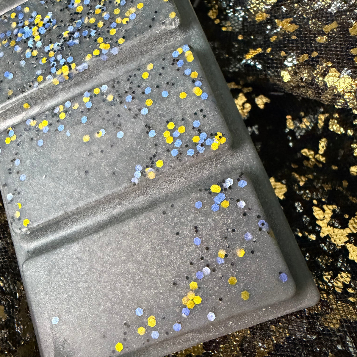 Detailed photograph of the Shadow Daddy wax melt showing gold circles, silver glitter and small iridescent pieces scattered across two charcoal-grey segments.