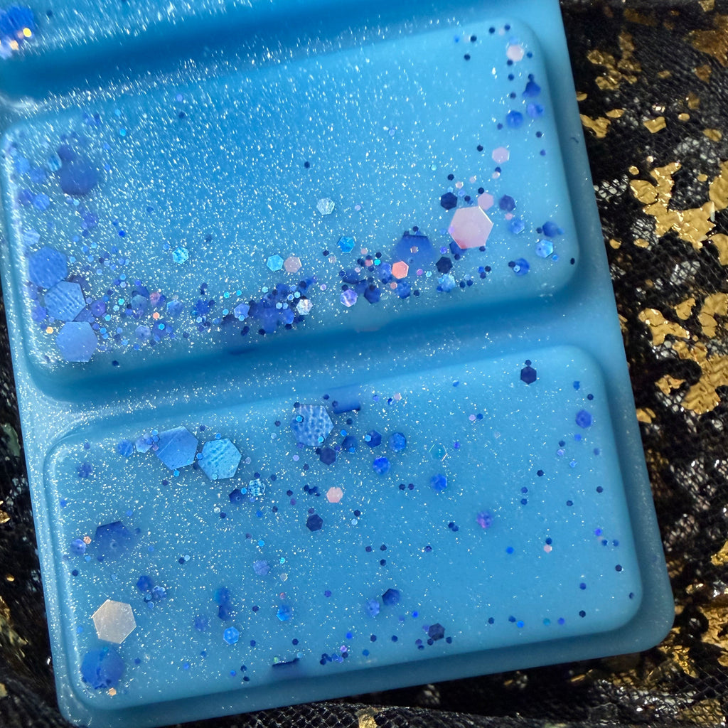 Close-up of the flat underside of The Snowy Escape wax melt showing vivid blue wax with scattered purple and silver glitter.