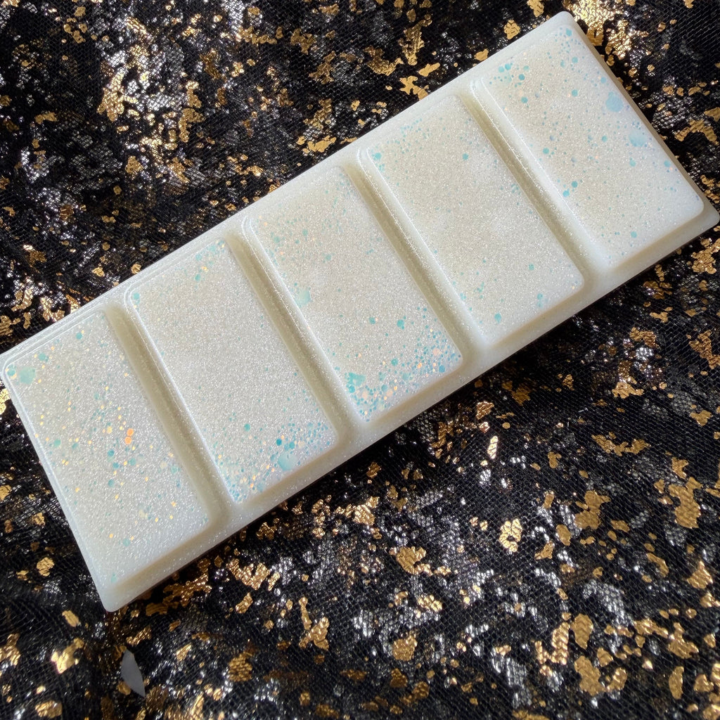 Unwrapped Xavier Acrux vegan soy wax melt positioned diagonally on black and gold fabric, with five ivory segments, pearly shimmer and pale blue and peach glitter.
