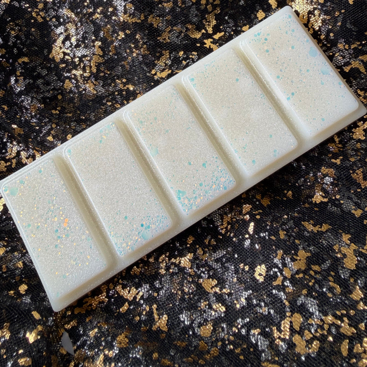 Unwrapped Xavier Acrux vegan soy wax melt positioned diagonally on black and gold fabric, with five ivory segments, pearly shimmer and pale blue and peach glitter.