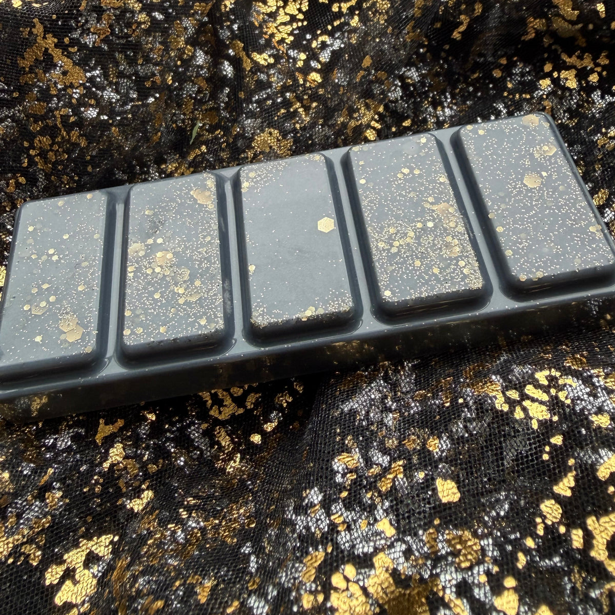 Side-angled photograph of the I Prefer the Villain five-segment charcoal wax melt, showing its raised rectangular sections and scattered gold glitter decoration.