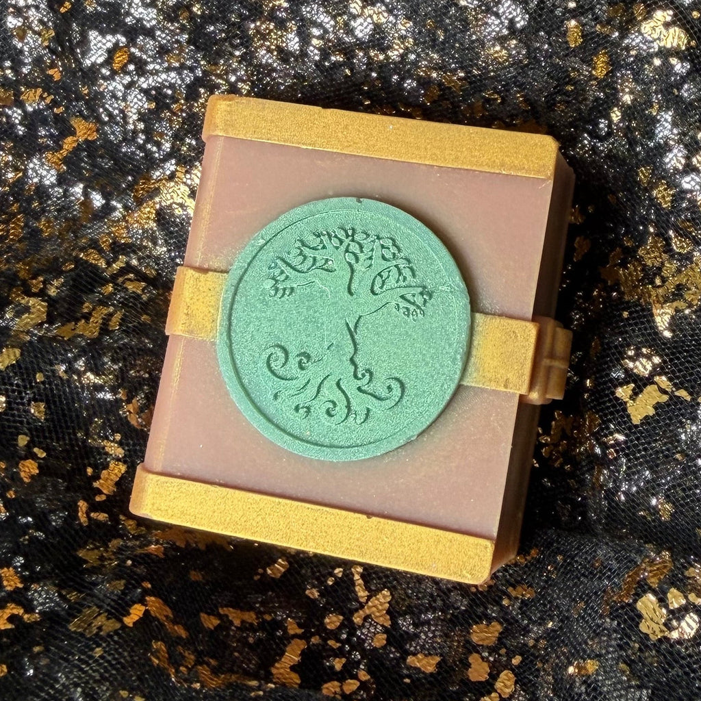 Side angled view of the Earth spellbook wax melt, with its mint tree emblem, gold clasp and right edge visible against black and gold fabric.