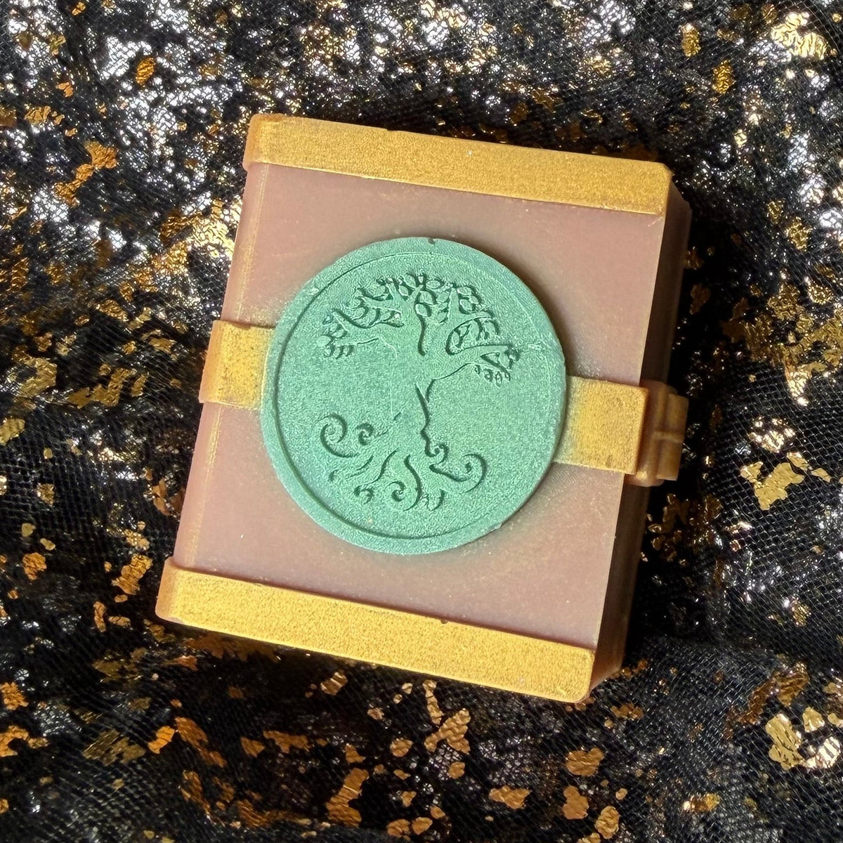 Side angled view of the Earth spellbook wax melt, with its mint tree emblem, gold clasp and right edge visible against black and gold fabric.