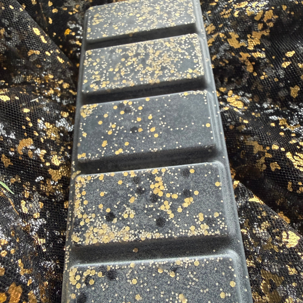 Zoomed-in view along the five rectangular segments of the Blonde Bombshell wax melt, showing charcoal-black wax with fine gold glitter, gold hexagons and small black circular decoration.
