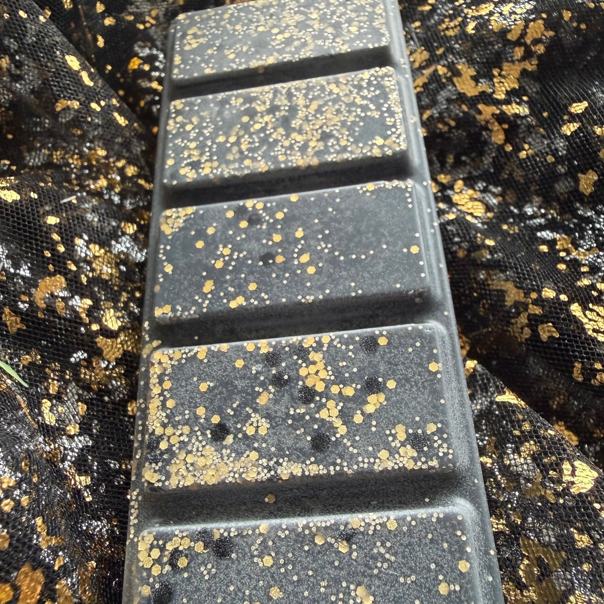 Zoomed-in view along the five rectangular segments of the Blonde Bombshell wax melt, showing charcoal-black wax with fine gold glitter, gold hexagons and small black circular decoration.
