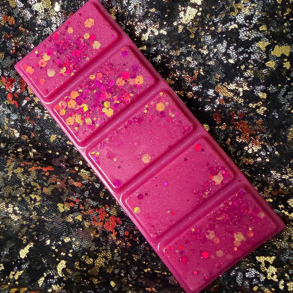 Full-length HMS Blossom wax melt positioned diagonally on black-and-gold fabric, showing all five vivid pink segments and gold glitter decoration.