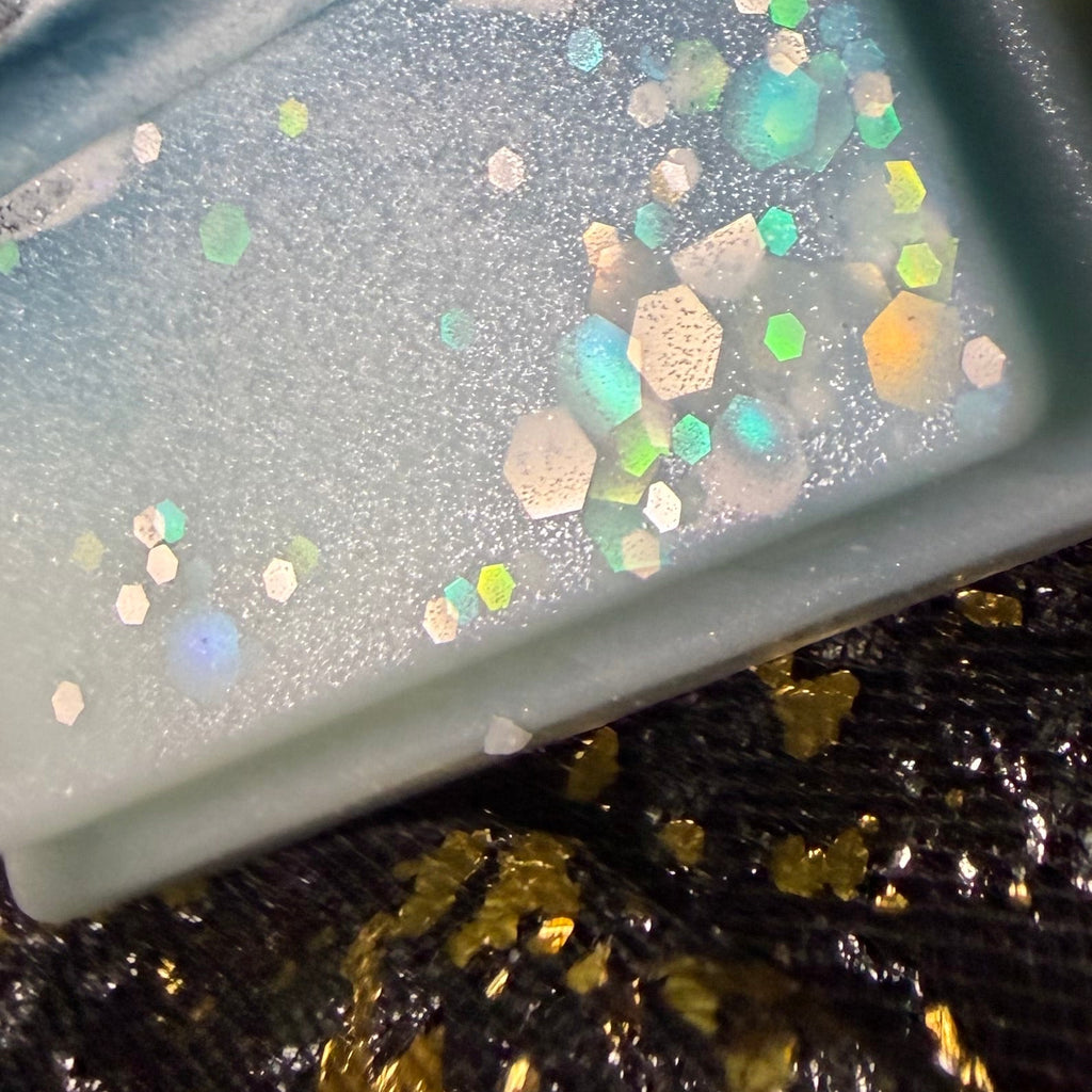 Zoomed-in photograph of the Lime Basil & Mandarin wax melt showing holographic glitter pieces reflecting green, blue, yellow and pink against the pale aqua wax.