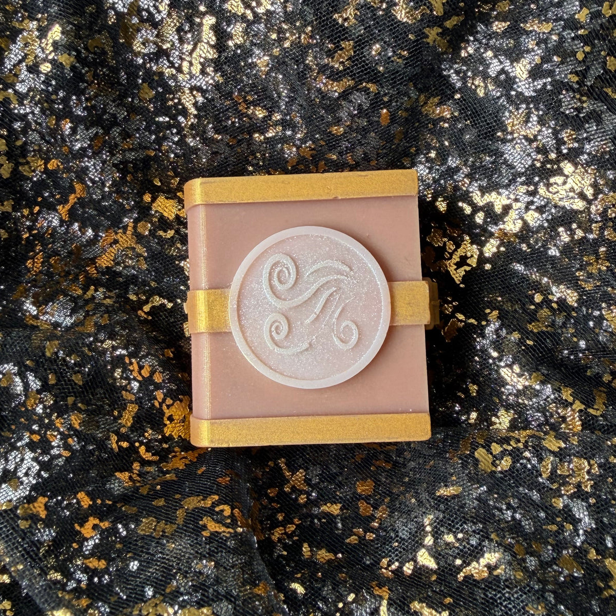 Air pine, eucalyptus and lavender scented spellbook wax melt, with a pale pearly wind swirl emblem and gold bands across its dusty rose cover.