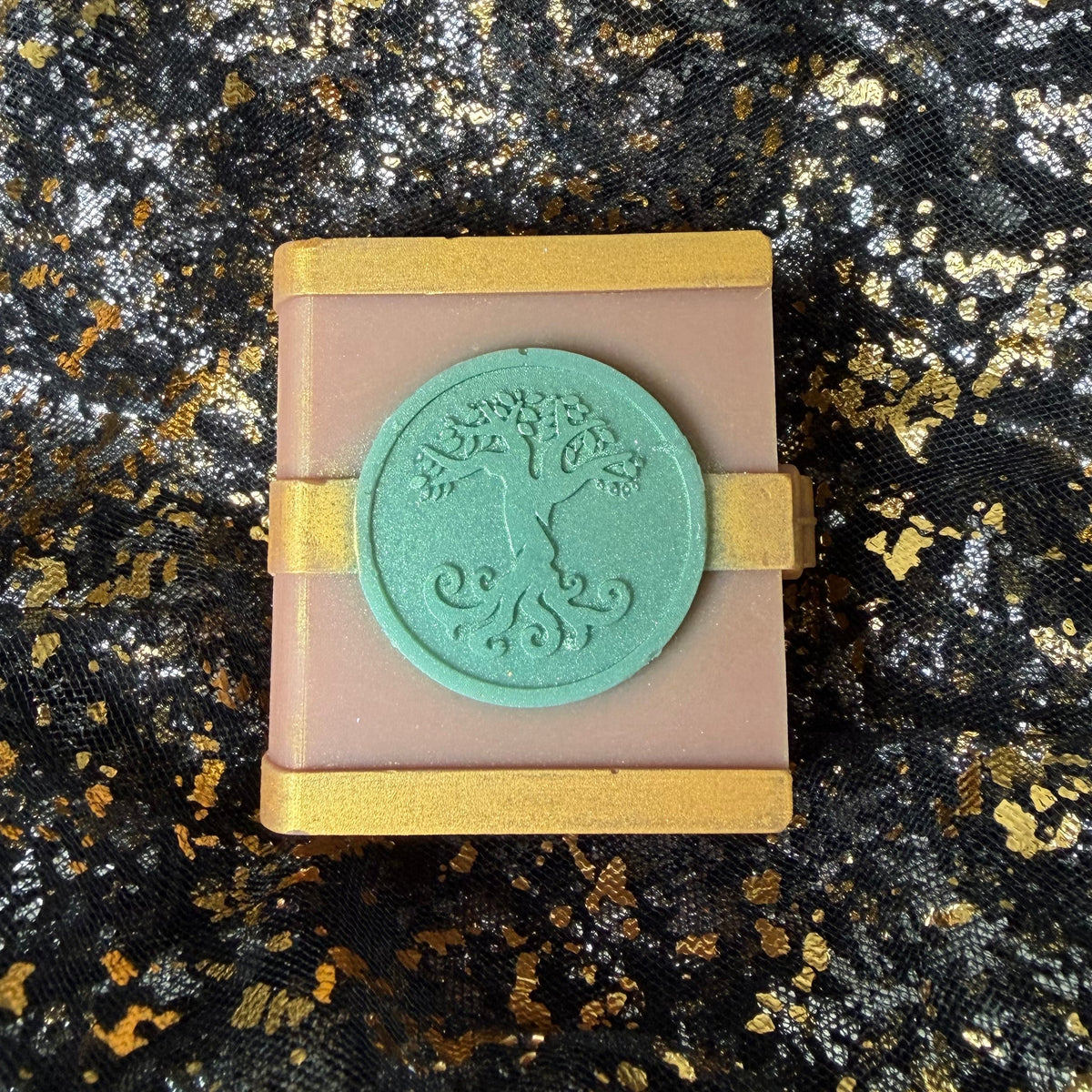 Full Earth soy wax spellbook centred on black and gold fabric, with a dusty rose cover, mint green tree emblem and gold detailing.
