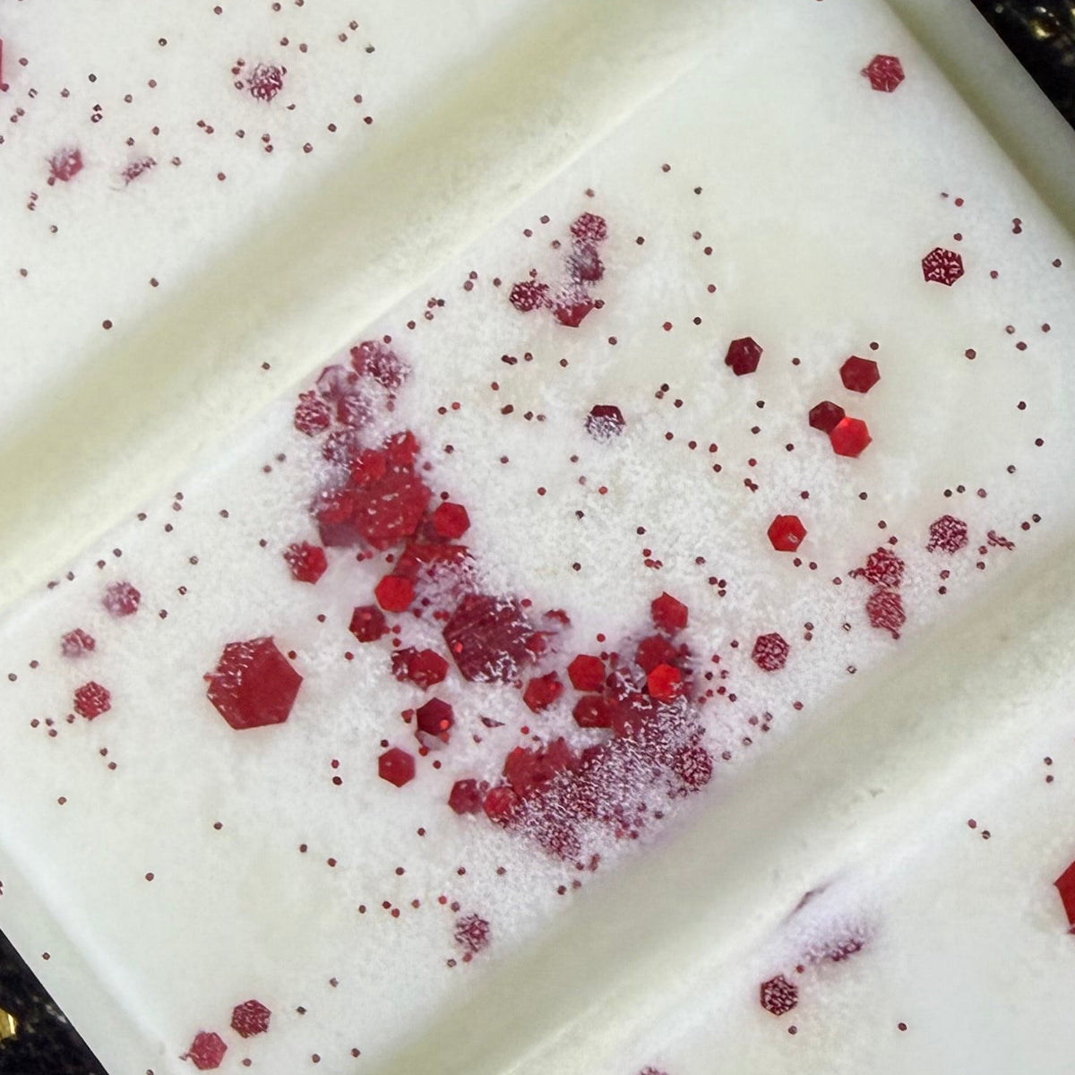 Close-up of the Forbidden Fruit wax melt showing fine red glitter, crushed red flakes and larger ruby-red hexagonal pieces scattered across the smooth ivory segments.