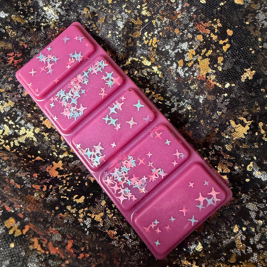Full-length One More Chapter wax melt positioned diagonally on black-and-gold fabric, showing all five deep pink segments and scattered star-shaped glitter.