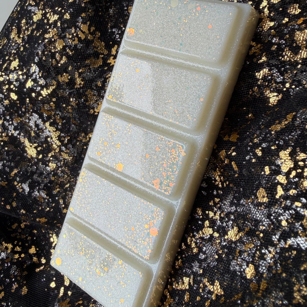 Side angled view of Xavier Acrux’s five segment lemongrass wax melt, with pearl white wax and peach gold glitter against black and gold fabric.
